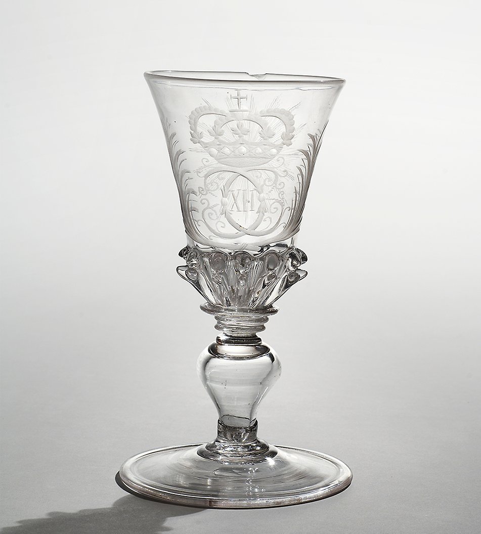 Vinglas på fot, utfört på Kungsholms Glasbruk i början av 1700-talet. På cuppans framsida finns Karl XII:s namnchiffer under kunglig krona och omgivet av palmkvistar, på motsatta sidan en strålande sol. Nästintill alla föremål från Kungsholms Glasbruk är idag drabbade av s.k. glassjuka, som resulterar i en mjölkaktig missfärgning. Likt delar av porslinsbeståndet, skingrades de äldre omoderna glasen i början av 1800-talet. Den nuvarande samlingen av Kungsholmsglas är resultatet av Oskar II:s samlargärning under 1800-talets slut.